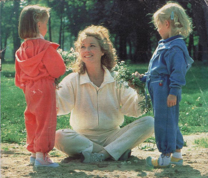 Woman sitting on grass holding flowers, smiling at young twins in red and blue outfits playing outdoors.