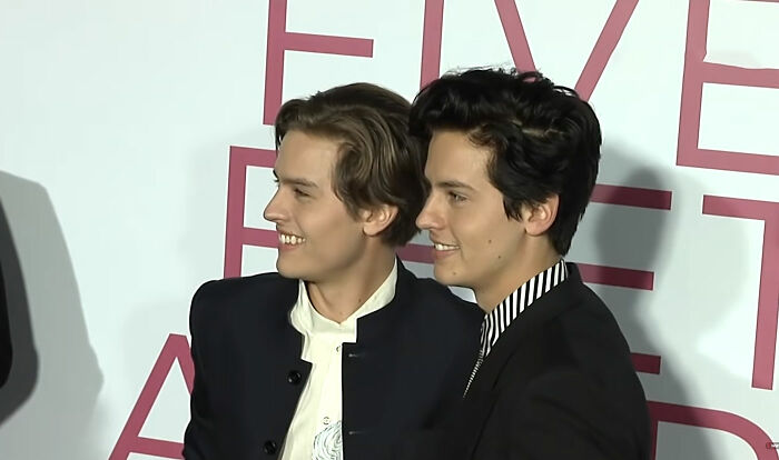 Celebrity twins smiling side by side at an event, dressed in formal black and white outfits, showcasing celebrity twins style.