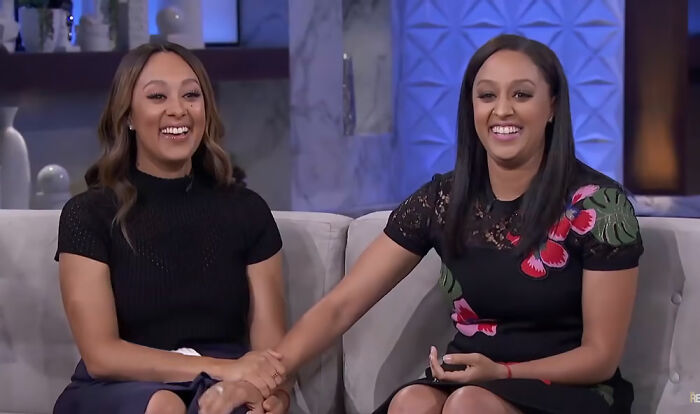 Two celebrity twins sitting on a couch smiling and holding hands during a TV interview about celebrity twins.