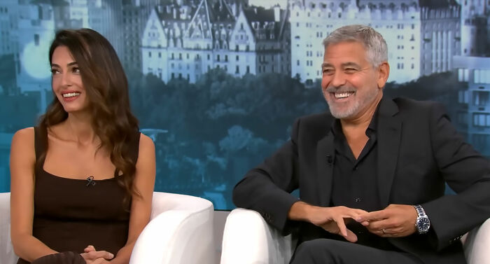 Celebrity twins sitting together in a television interview, smiling and engaged in a friendly conversation.