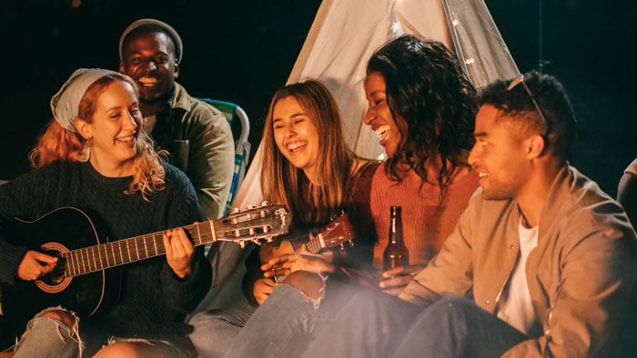 Millennials enjoying a campfire, playing guitar and laughing, in a casual outdoor setting.