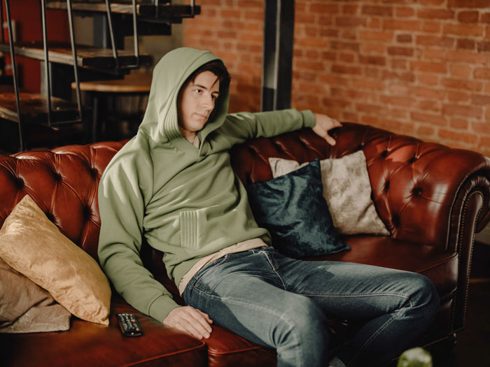 Millennial relaxing on a red leather couch in a green hoodie, surrounded by pillows in a cozy, casual setting.