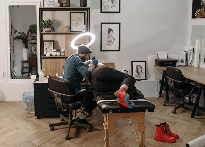 Tattoo artist working in a studio, tattooing a footprint on a client's arm, surrounded by art and equipment.
