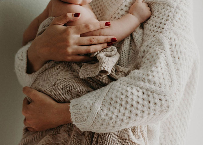 New mom holding baby in soft knit sweater, discussing MIL wanting tattoo of grandkid's footprint.