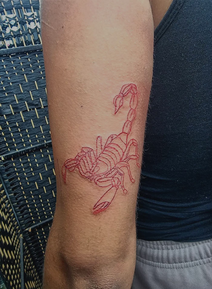 Red ink tattoo of a detailed scorpion on a person's arm against a wicker background.