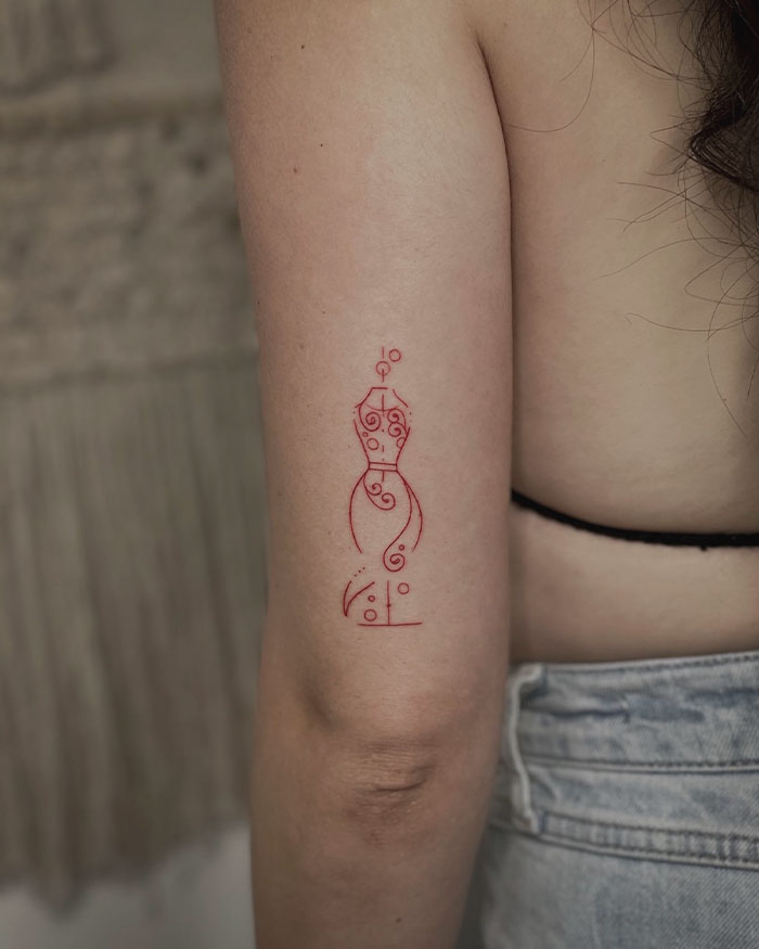 Simple red ink tattoo of an abstract dress form on a person's upper arm.