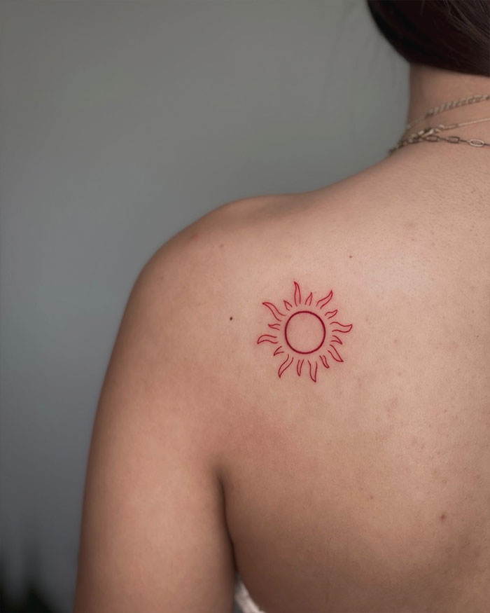 Red ink tattoo of a minimal sun design on a person's shoulder.