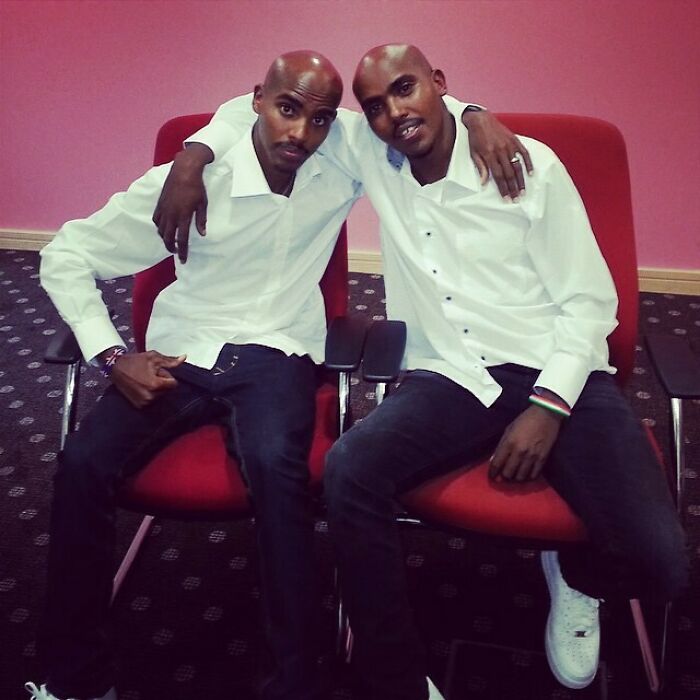 Two celebrity twins seated on red chairs, wearing white shirts and dark pants, posing together indoors.