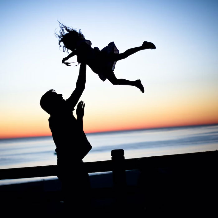 Silhouetted older person lifting child in the air at sunset by the ocean, sharing advice for younger people.