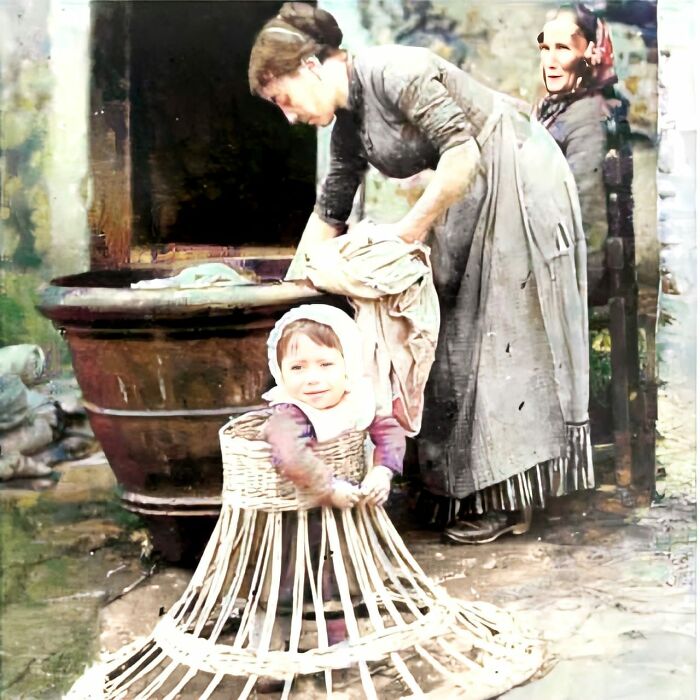 Un bebé aprende a andar en un marco de mimbre mientras su madre lava la ropa. Alrededor de 1910