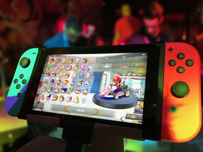 Nintendo Switch displaying Mario Kart character selection with vibrant red and blue controllers in a colorful gaming environment.