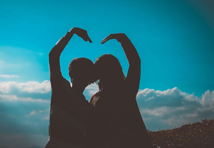 Silhouettes of two people over 50 forming a heart shape with arms against a bright blue sky with clouds.