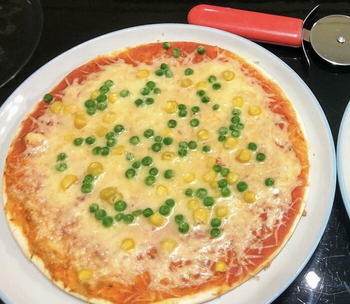 Unappetizing pizza topped with scattered peas and corn, on a white plate with a red-handled pizza cutter nearby.