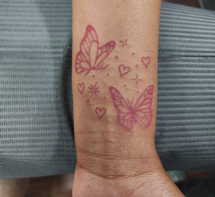 Red ink tattoo of butterflies and hearts on a person's wrist.