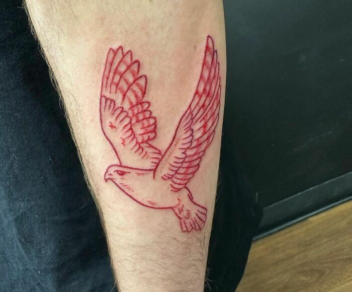 Red ink tattoo of a flying bird on a person's forearm.