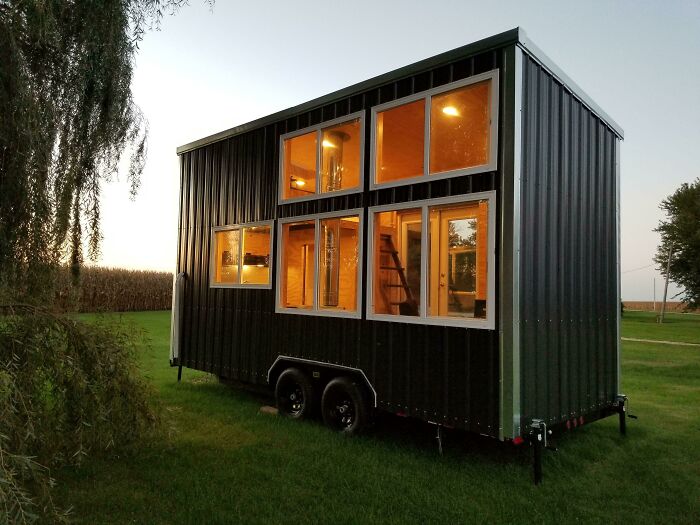 Modern tiny house on wheels with large windows, set against a serene grassy landscape at dusk.