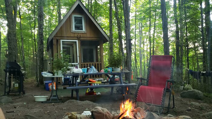 Cozy tiny house in a forest setting with a campfire, picnic table, and a red chair, showcasing irresistible tiny house designs.