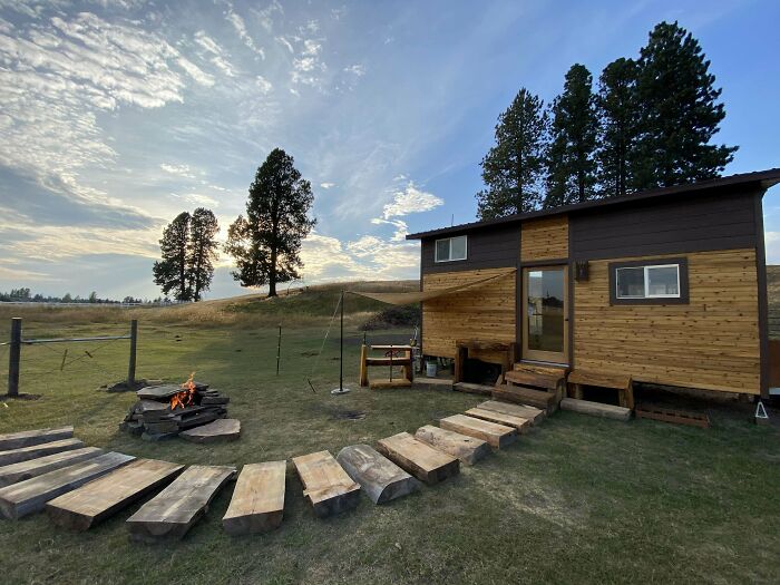 Tiny house design with a rustic wooden exterior, set in a scenic landscape with a stone fire pit and wooden path under a blue sky.