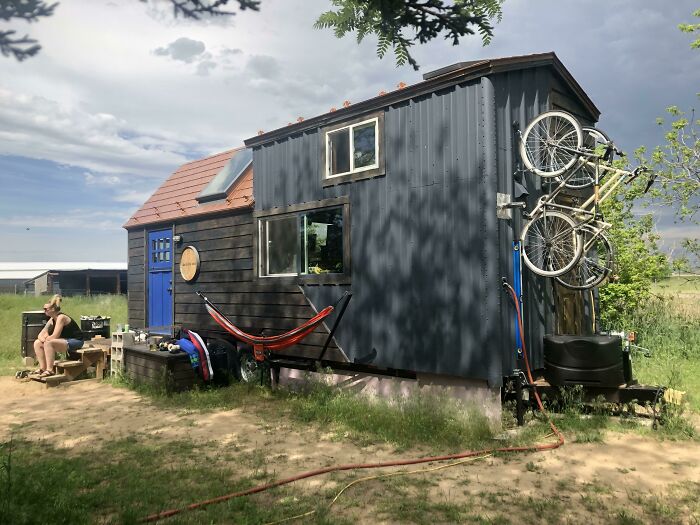 Black tiny house with bicycles mounted on the wall, surrounded by grass and a person sitting nearby enjoying the outdoors.