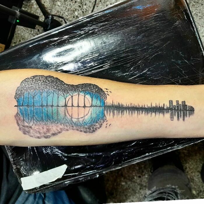 Music tattoo of a guitar reflecting on water with sound waves and cityscape design on arm.