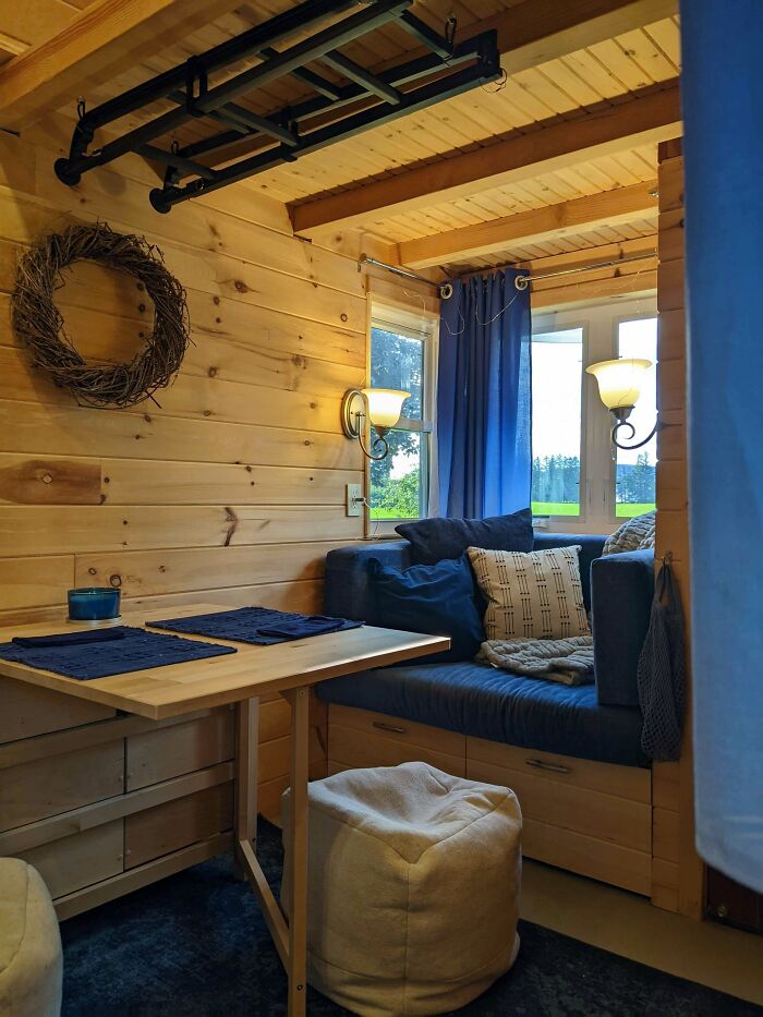 Cozy wooden tiny house interior with a blue couch, dining table, and natural light coming through windows.
