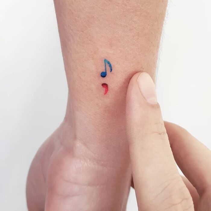 Small music tattoo with a blue note and red semicolon on wrist.