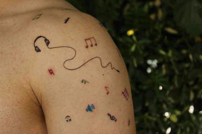 Music tattoos featuring notes, a treble clef, headphones, and colorful symbols on a person's shoulder against a leafy background.