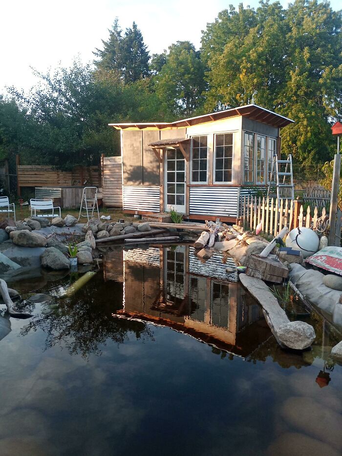 Tiny house design with a pond, surrounded by trees and garden chairs, reflecting a serene natural setting.