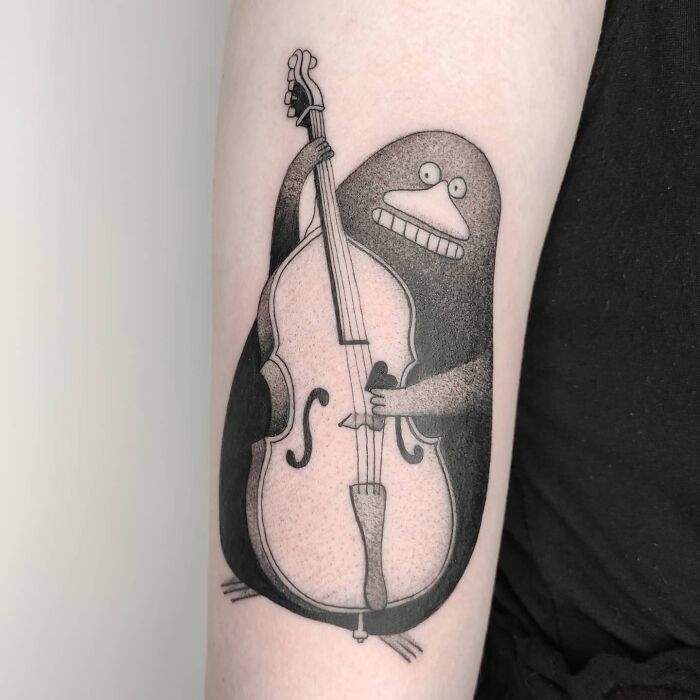 Music tattoo featuring a whimsical creature playing a double bass, inked on a person's arm.