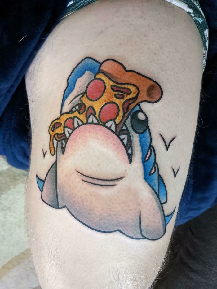 Food tattoo of a cartoon shark eating a slice of pizza, inked on a person's leg.