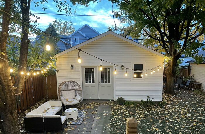 White tiny house with string lights hanging between trees over an outdoor seating area in a cozy backyard setting.