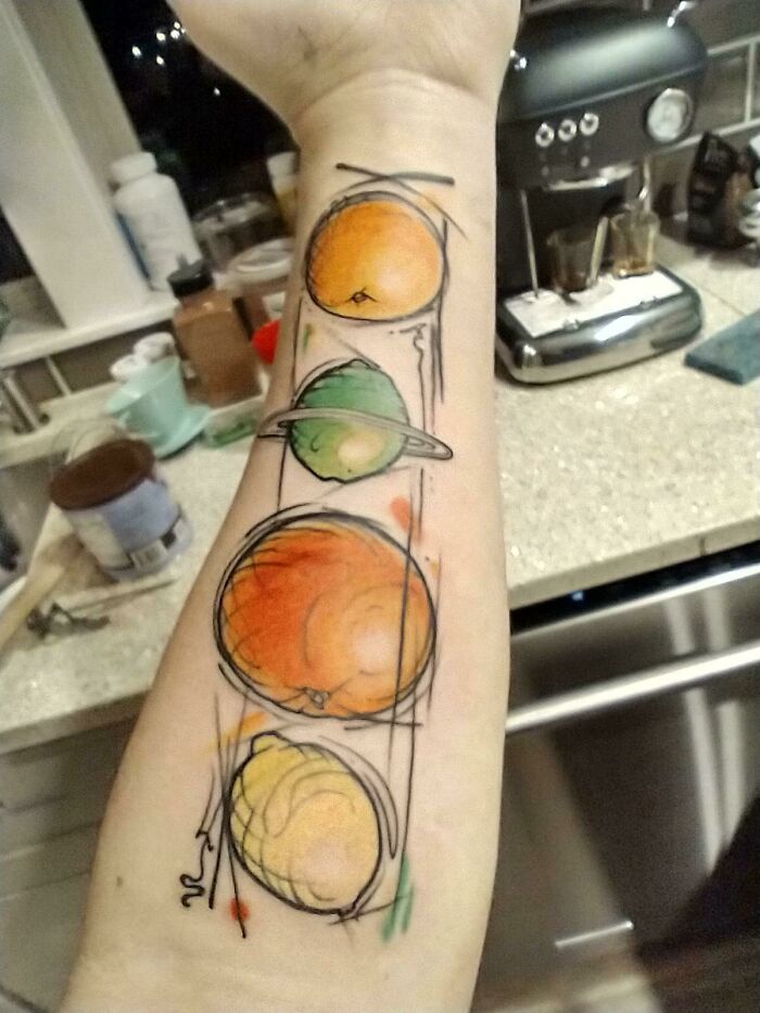 Colorful fruit-themed food tattoo on a forearm with kitchen background.