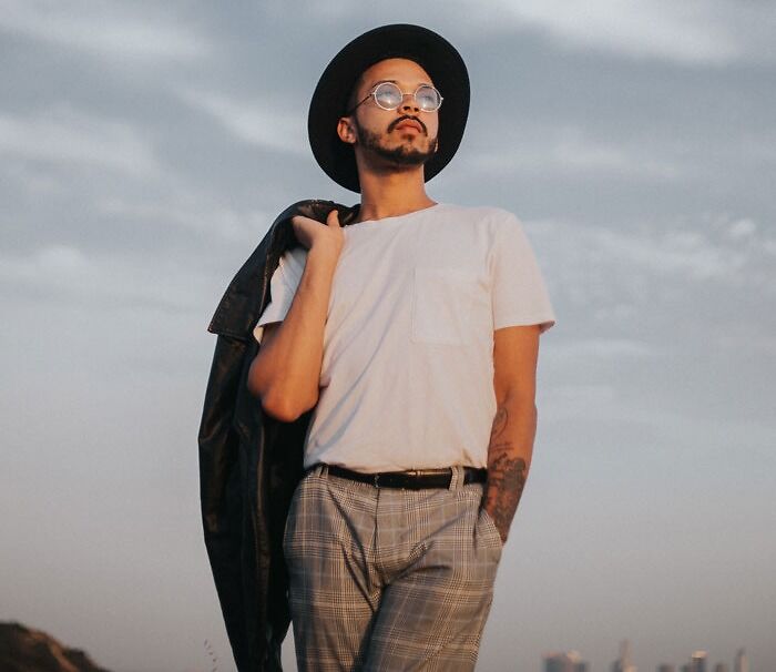 Man standing and wearing white shirt, patterned brown jeans and hat