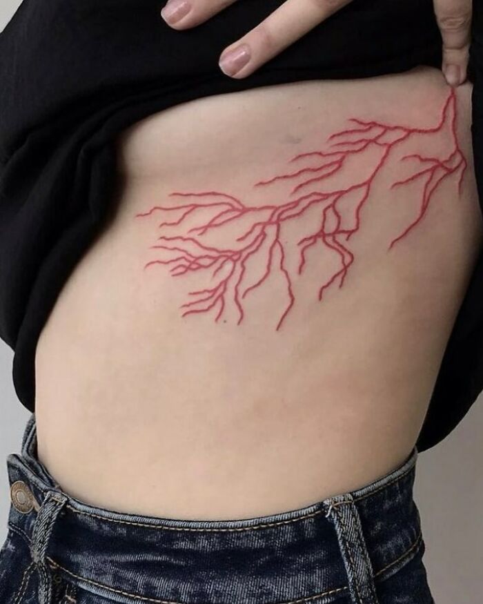 Intricate red ink tattoo on skin, resembling branching veins, showcased on the side torso above denim jeans.