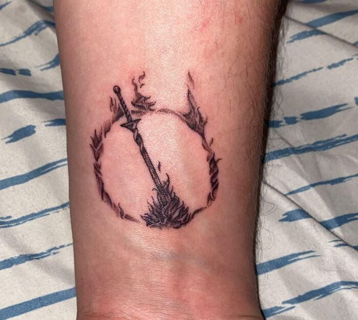 Wrist tattoo of a flaming circle with a sword in the center on a person's arm.