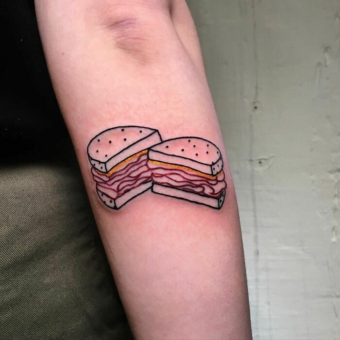 Food tattoo of a sliced burger on a forearm, featuring layers of bacon and cheese.