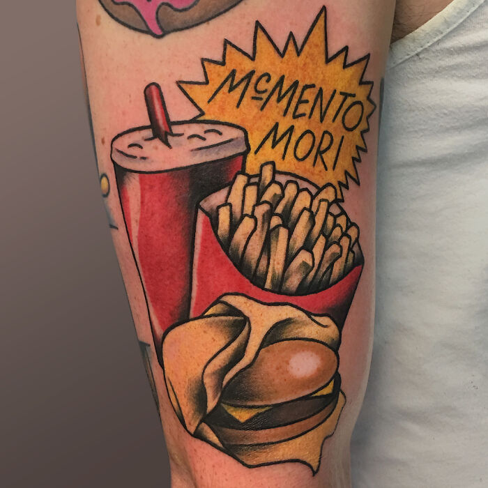 Watercolor food tattoo of a burger, fries, and cola with "memento mori" text on skin.