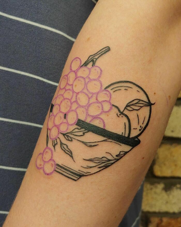 Food tattoo featuring a bowl of fruit, including grapes and an apple, on the arm.