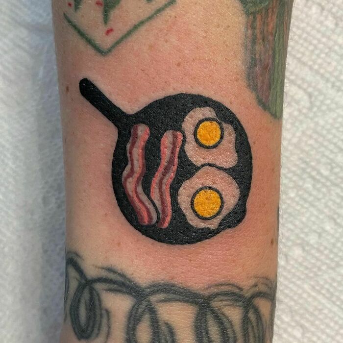 Tattoo of a frying pan with bacon and eggs, showcasing a delicious food-inspired design on the arm.