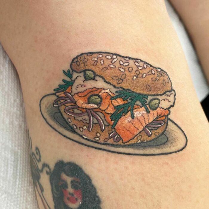 Food tattoo of a salmon bagel with cream cheese, onions, capers, and dill on a forearm.