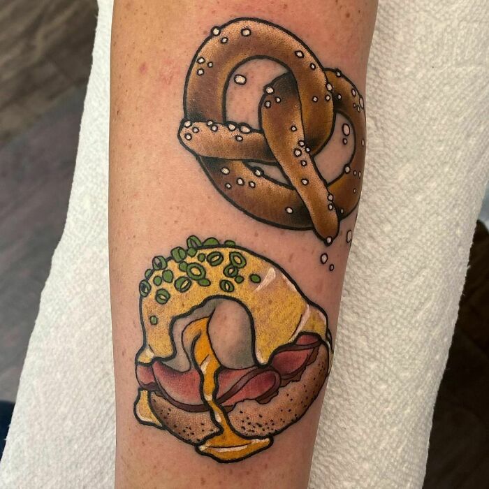 Food tattoos featuring a pretzel and a bacon-topped bagel on a person's forearm.