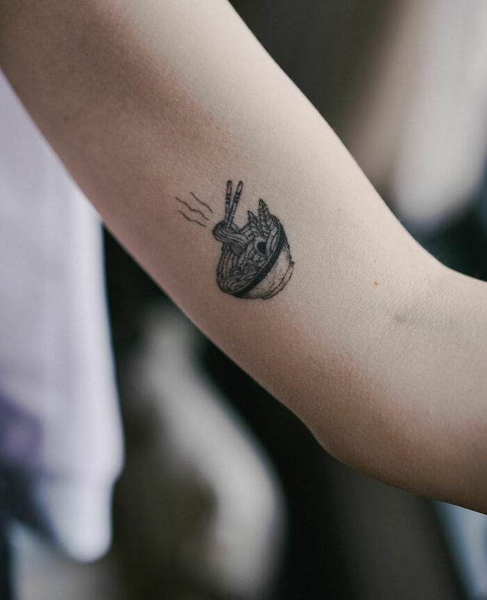 Food tattoo of steaming ramen bowl with chopsticks on forearm.