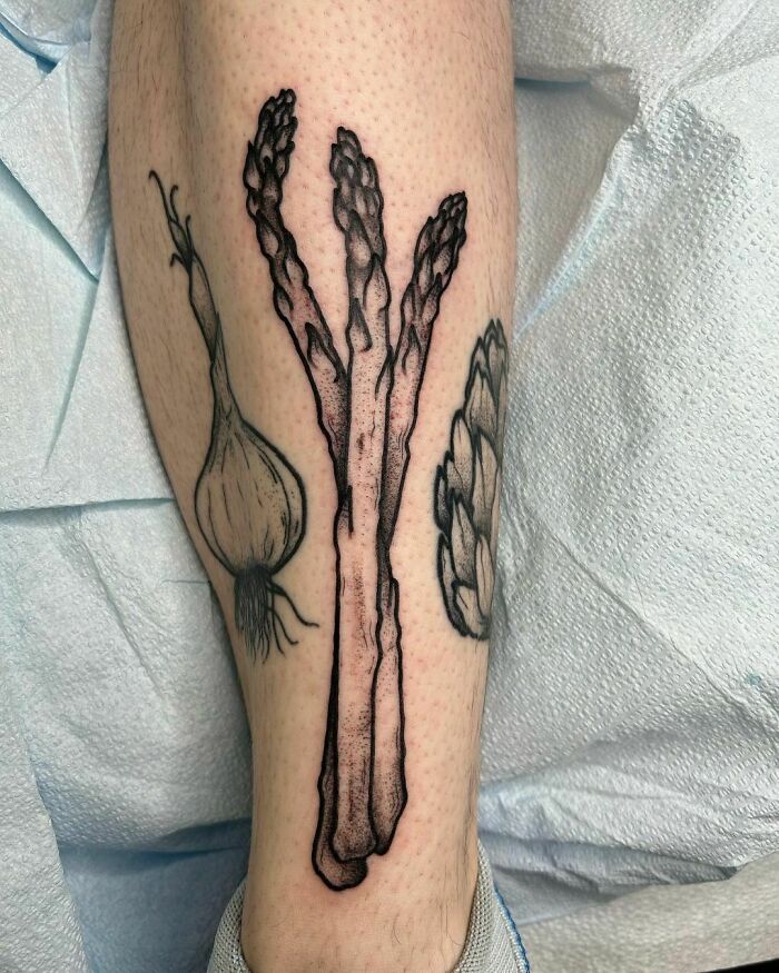 Tattoo of asparagus, onion, and artichoke on a leg, showcasing delicious food-themed ink artistry.
