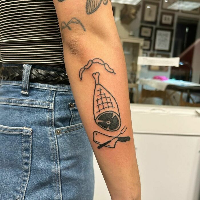 Black and white ham tattoo on arm, featuring a cleaver and fork design with geometric details.