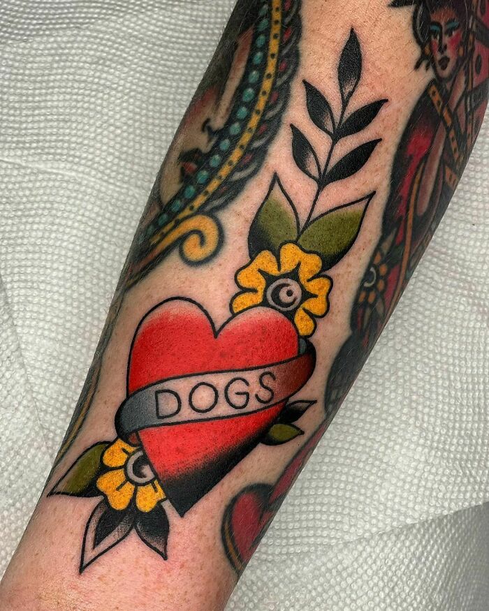 American traditional tattoo of a heart with "Dogs" banner and floral elements on the arm.