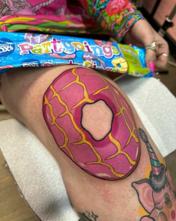Close-up of a vibrant food tattoo featuring a pink and yellow donut design on a knee, capturing creative and delicious ink art.