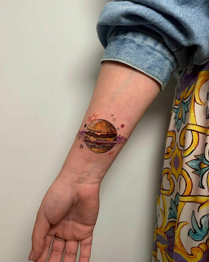 Colorful burger tattoo on arm, surrounded by stars, showcasing delicious food-themed ink art.
