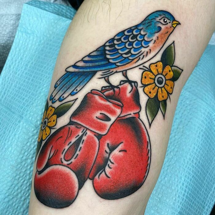 American traditional tattoo featuring a colorful bird, red boxing gloves, and yellow flowers on skin.