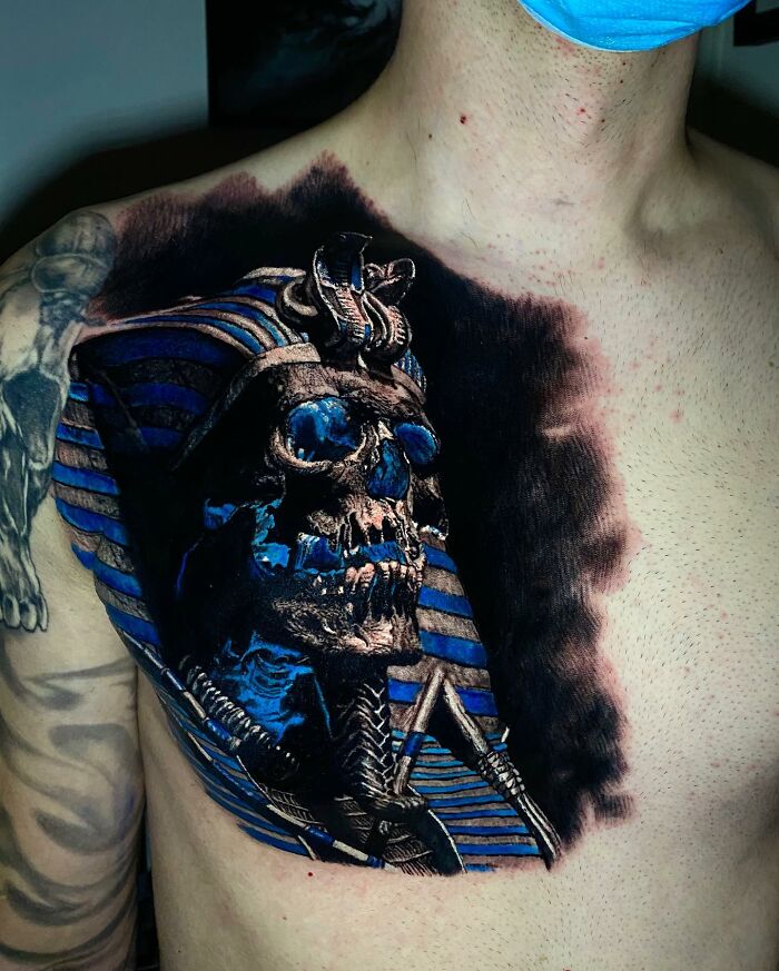 Tattoo art of a pharaoh skull with vivid blue accents on a person's chest, showcasing intricate and realistic design.