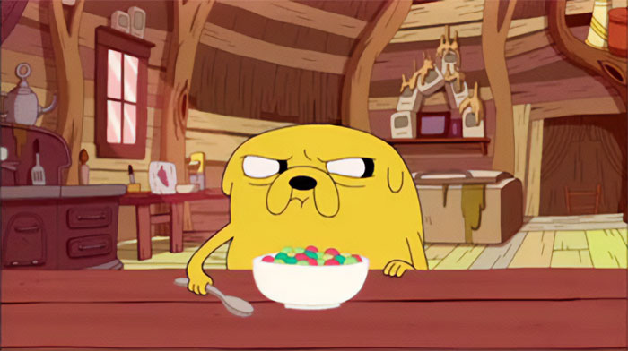 Jake the Dog from Adventure Time sitting at a table eating colorful corn flakes in a rustic wooden room setting.